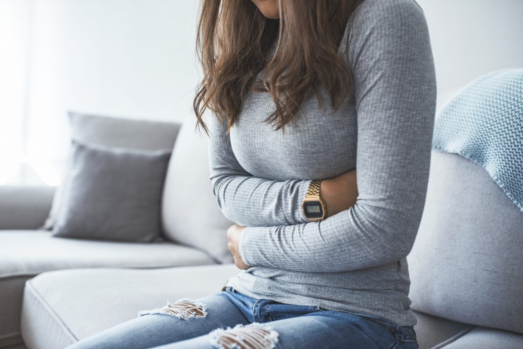 Woman sitting on couch holding her stomach as she feels stomach pain