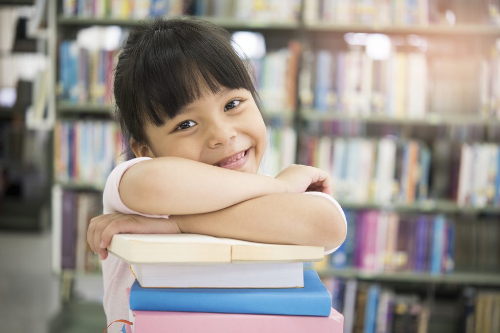 Girl smiling into the camera with her arms crossed across a stack of books.