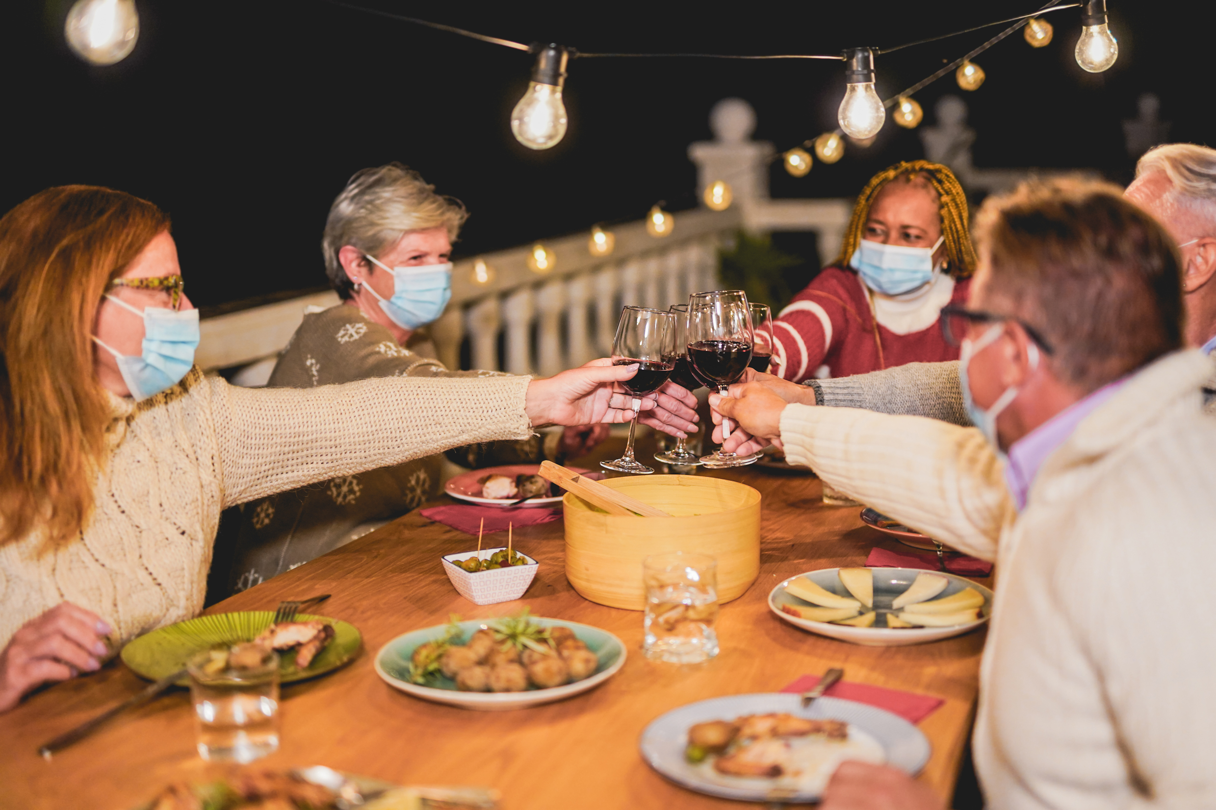 Happy multiracial senior people cheering with red wine at dinner while wearing surgical face mask for coronavirus