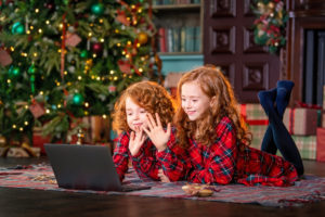 Red-haired children next to the Christmas tree and gifts communicate online through a laptop.