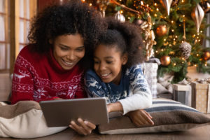 Jeune fille et sa mère regardent une tablette devant un sapin de Noël entouré de cadeaux.