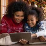 Close up happy African American woman with daughter using tablet together, lying on soft pillows near Christmas tree at home, happy family shopping online, choosing gifts, enjoying winter holiday