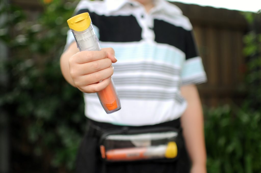Boy holding an epinephrine auto-injector, and a second auto-injector is shown in a pouch that is across his waist.
