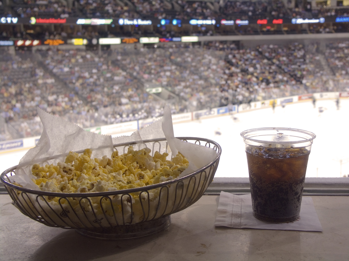 A bowl of popcorn with a glass of soda at a sports game.