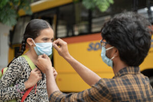 Father helping daughter to wear mask before getting inside the school bus as coronavirus or covid-19 safety measures - concept of back to school and new normal lifestyle.