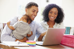 Parents With Baby Working In Office At Home Looking At Laptop Smiling